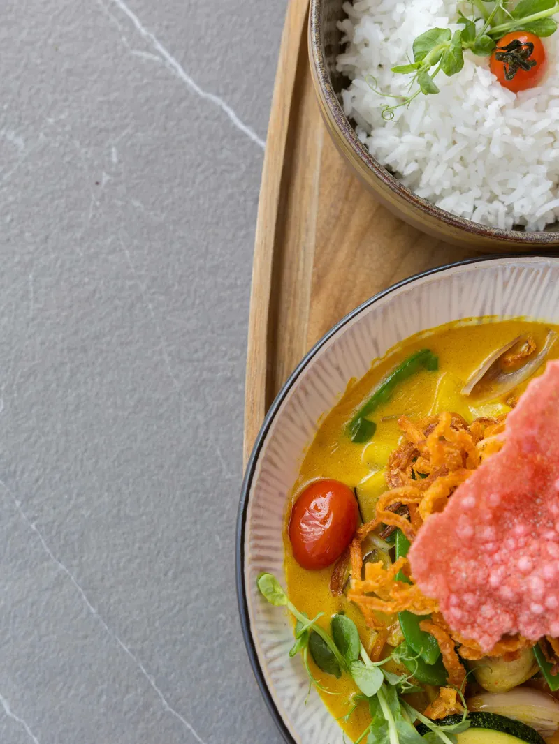 Gelbes Curry mit Gemüse, Cherrytomate und Krupuk neben einer Schale Reis