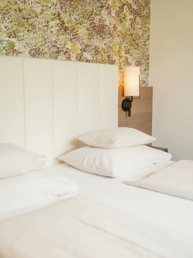 Bett mit weißen Kissen und Wandleuchte vor geblümter Tapete im Hotelzimmer