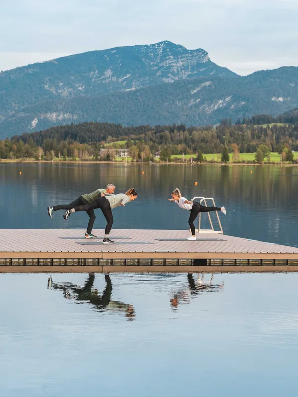 Personen machen Yoga auf einem Steg am See vor Bergkulisse