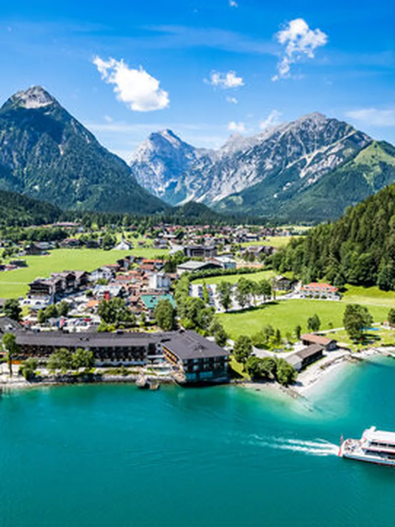 Malerischer Achensee mit türkisem Wasser, umgeben von grünen Bergen und einem Dorf am Ufer. Ein Passagierschiff fährt auf dem See.