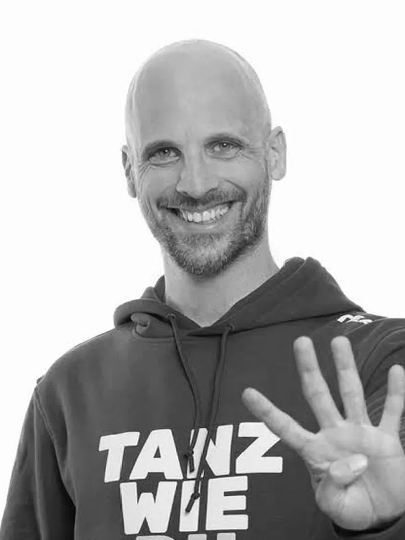Schwarz-Weiß-Foto eines lächelnden Mannes mit Glatze und Bart, in einem "TANZ WIE" Hoodie, der vier Finger zeigt.