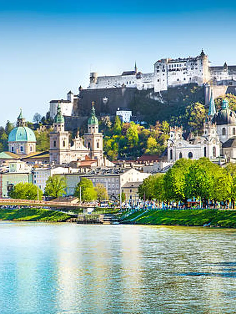 Salzburg: Festung Hohensalzburg thront über der Altstadt am Fluss Salzach unter blauem Himmel mit grünen Bäumen.