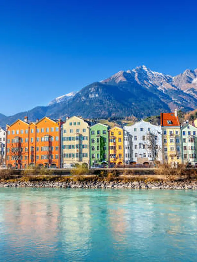 Farbenfrohe Häuserreihe am Inn-Fluss in Innsbruck, mit schneebedeckten Alpen im Hintergrund unter blauem Himmel.
