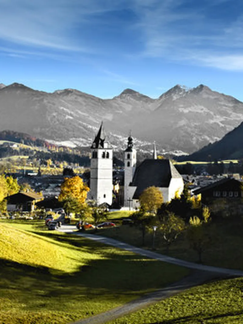 Alpenlandschaft im Herbst: Eine weiße Kirche im Tal, umgeben von goldenen Bäumen und grünen Hügeln, vor majestätischen Bergen und blauem Himmel.