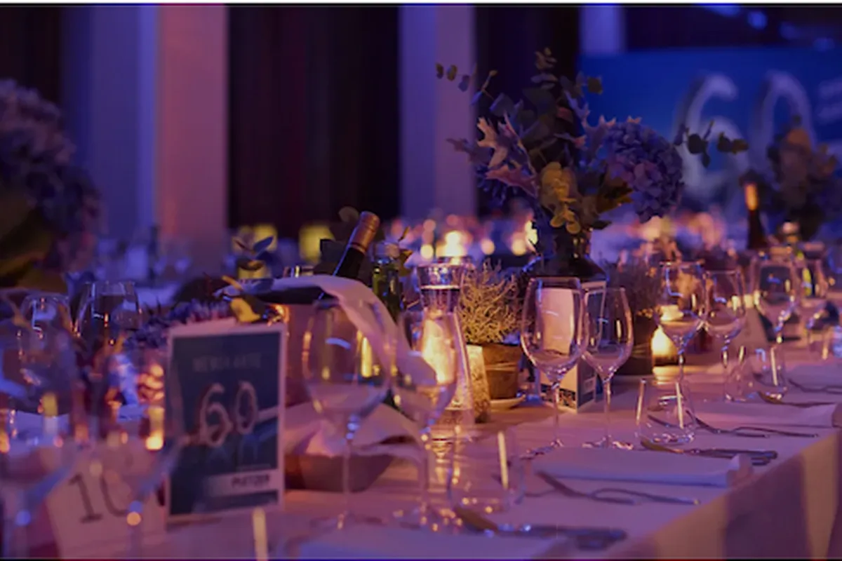 Festlich gedeckte lange Tafel mit Gläsern, Blumen, Kerzen und der Zahl 60, beleuchtet in Blau und Lila.