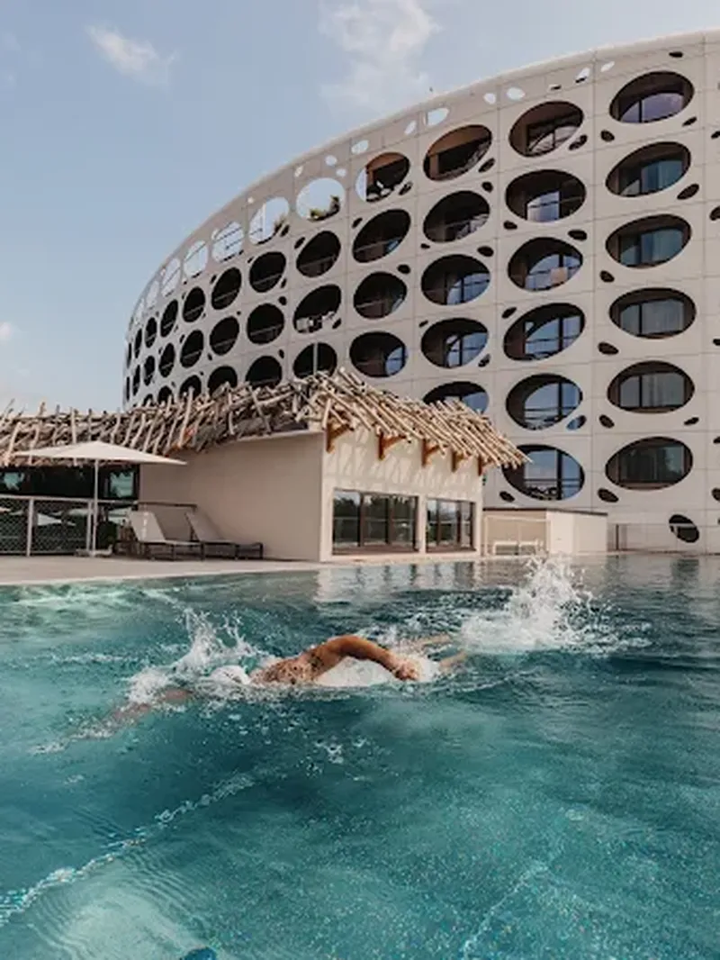 Eine Person schwimmt in einem Infinity-Pool vor einem modernen, geschwungenen Gebäude mit ovalen Fenstern und einem rustikalen Holzhaus.