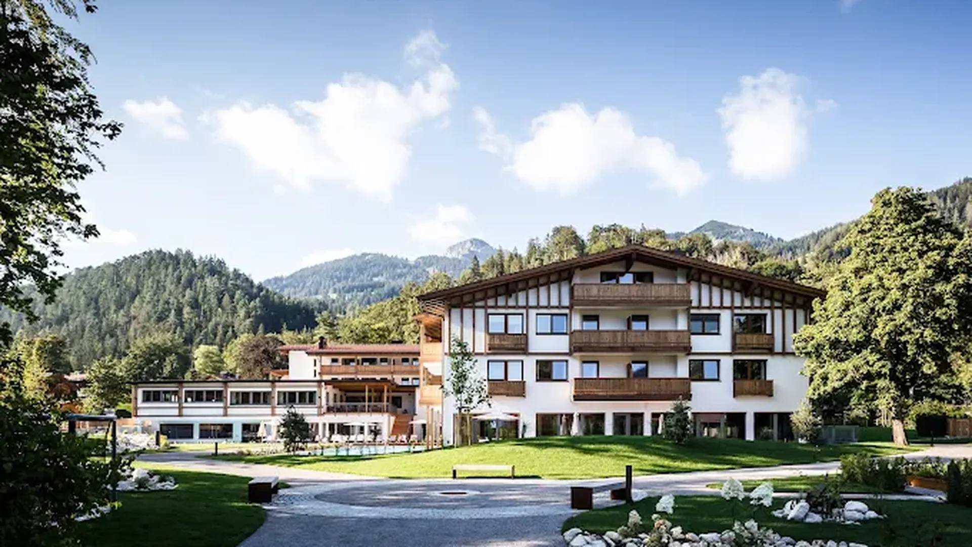 Großes Hotel im alpenländischen Stil, umgeben von grünen Wiesen und Bäumen, mit bewaldeten Bergen unter blauem Himmel im Hintergrund.