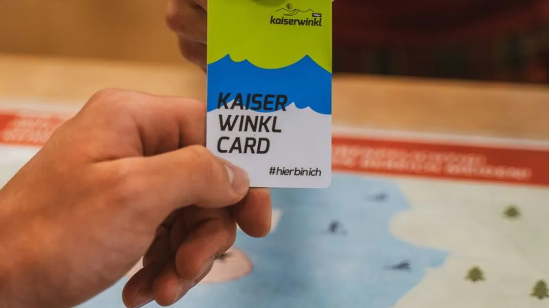 Eine Hand übergibt eine grüne, blau-weiße "Kaiserwinkl Card" an eine andere Hand, über einer Landkarte.