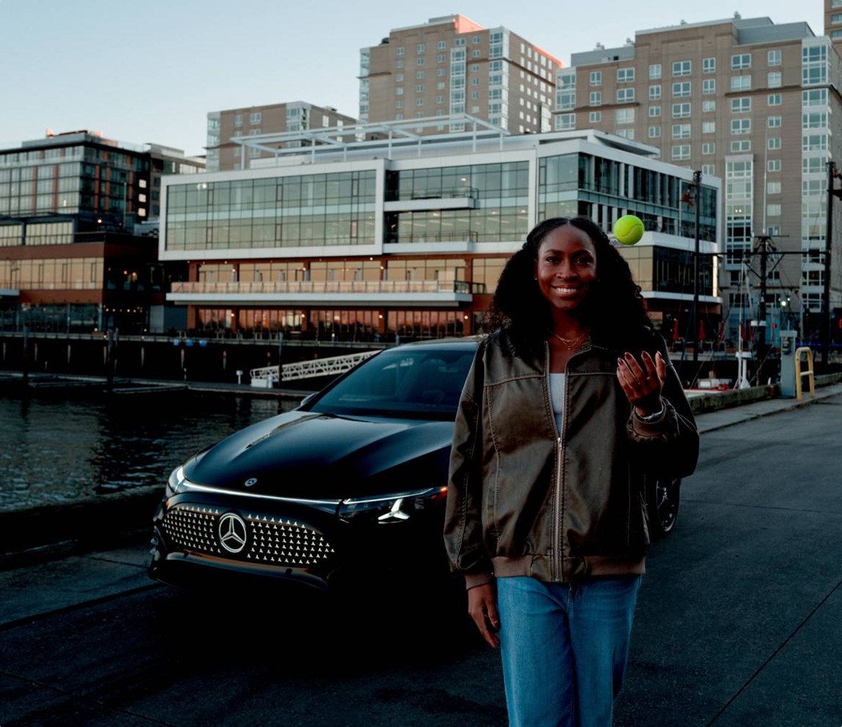 Jeune femme souriante lançant une balle de tennis devant une Mercedes-Benz noire, au bord de l'eau avec des bâtiments urbains en arrière-plan.