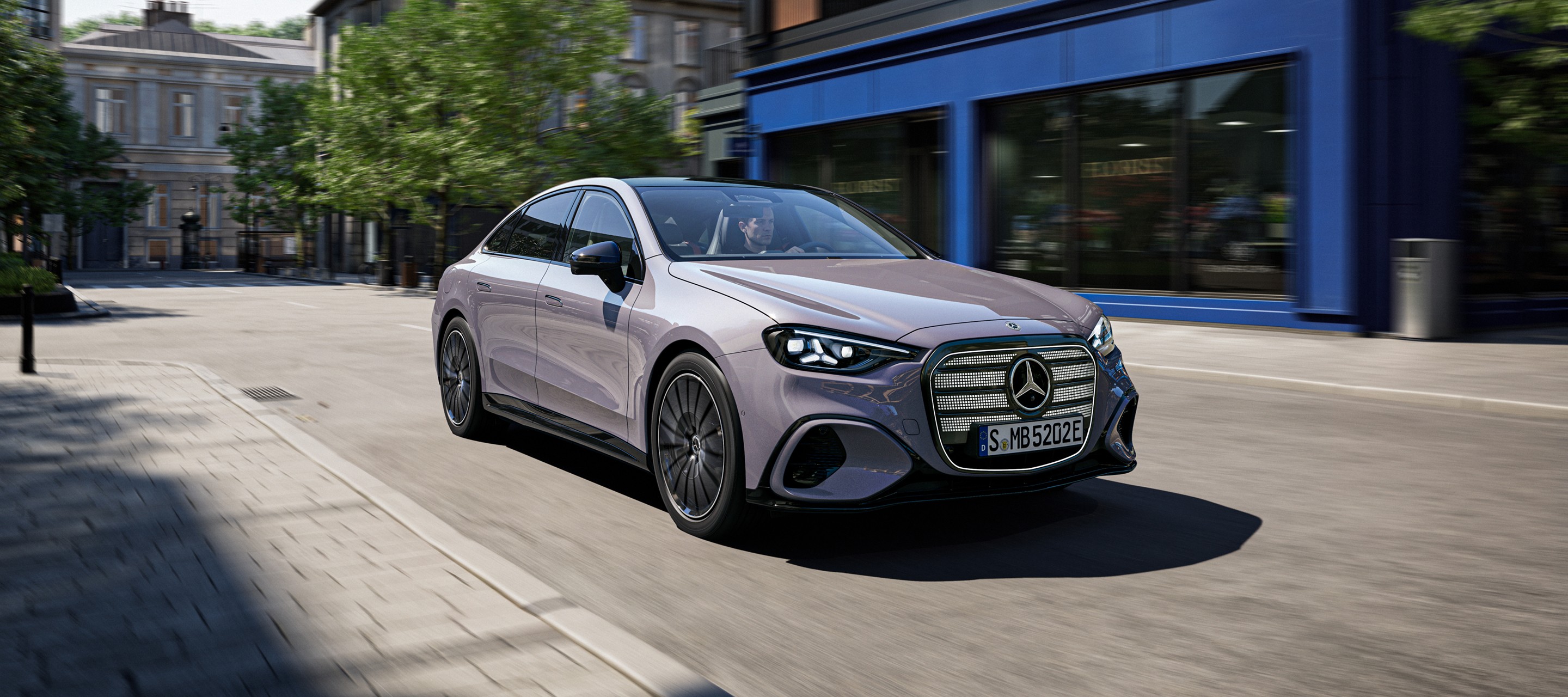 Mercedes-Benz Classe E grise roulant sur une avenue urbaine, vue trois quarts avant, avec bâtiments et arbres en arrière-plan.