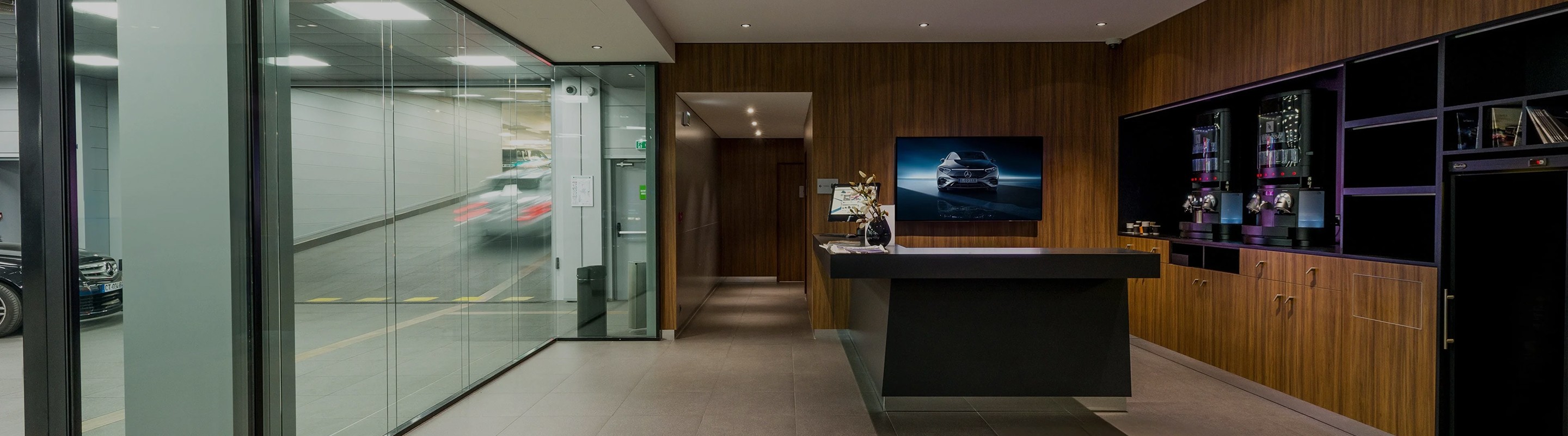 Intérieur moderne d'une concession automobile avec un comptoir sombre, des murs en bois et une cloison vitrée donnant sur un parking.