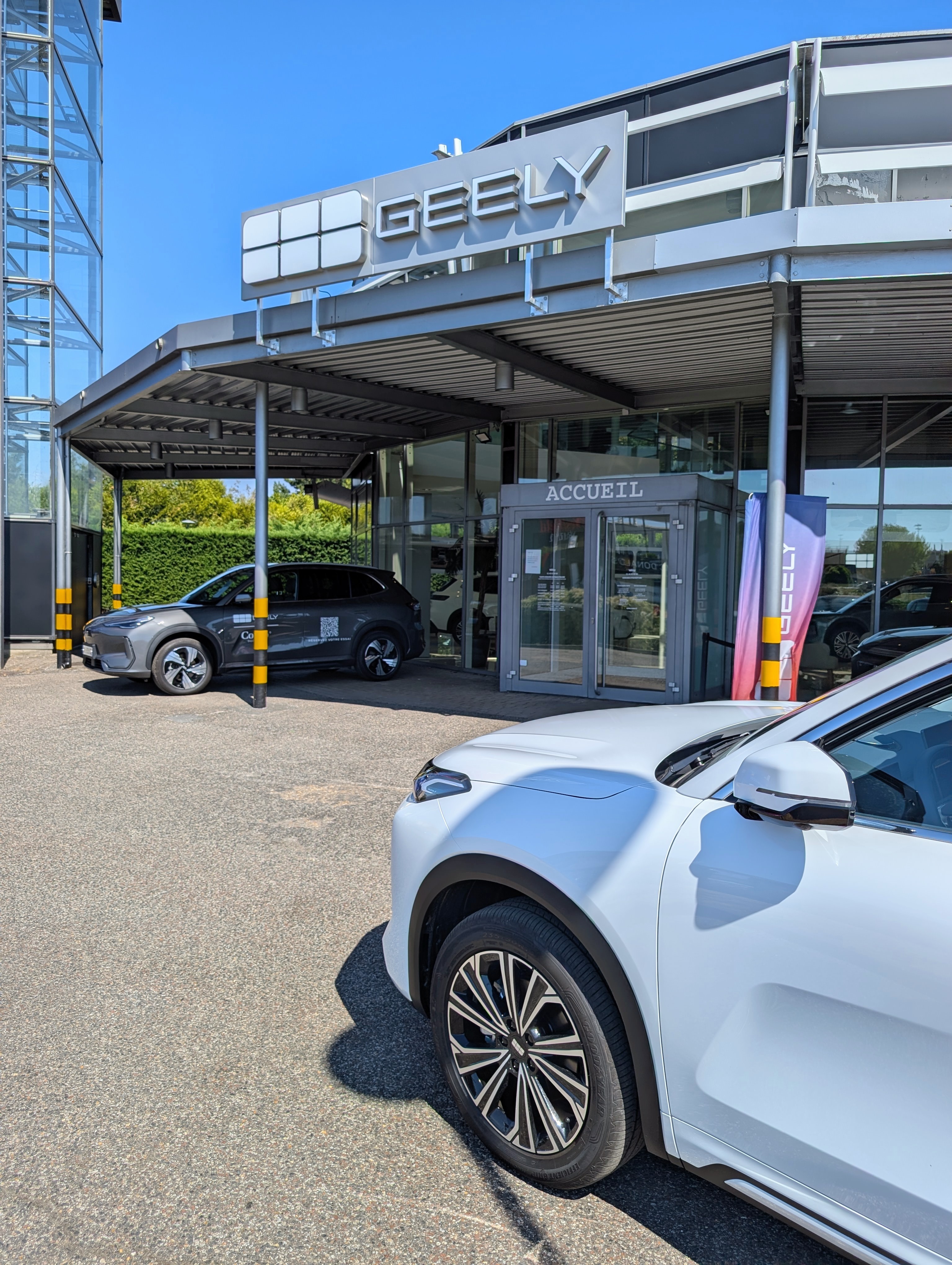 Entrée d'une concession Geely avec deux véhicules garés devant, dont un SUV blanc au premier plan, par temps ensoleillé.