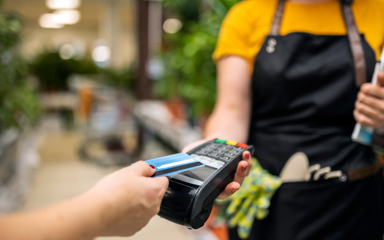 A person wearing an apron holds a card reader as another swipes a blue credit card, navigating not just purchases but unseen exchange rates. The background reveals a store with blurred shelves and greenery.