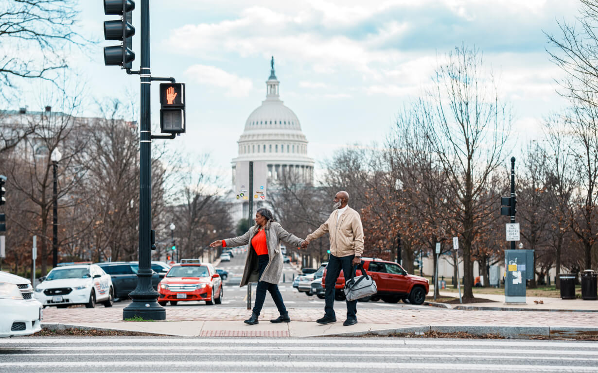A couple holding hands crosses a street near the United States Capitol. The sky is cloudy, and trees line the street. Traffic lights are visible, and cars are passing by in the background.