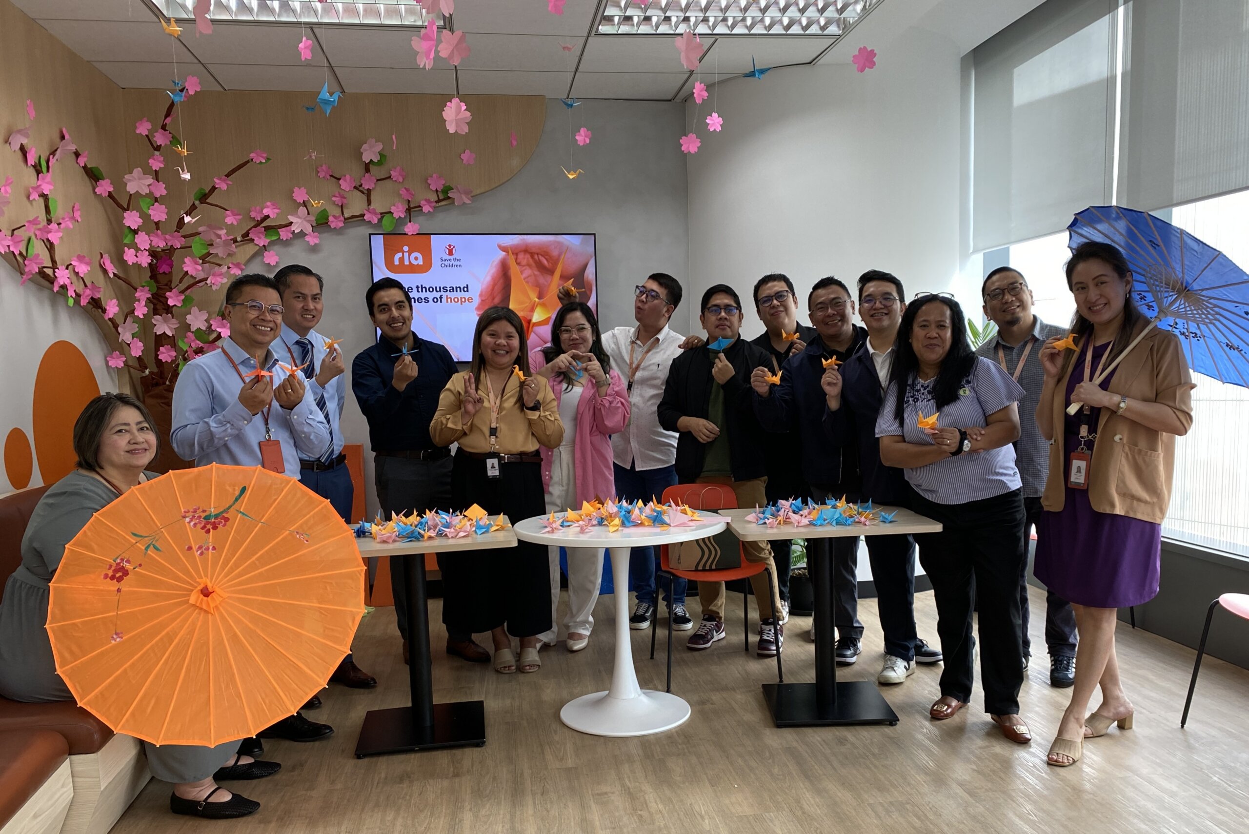 A group of people stand smiling in an office decorated with pink paper flowers and origami cranes, holding colorful umbrellas and origami pieces. A screen and tables with more origami are in the background.