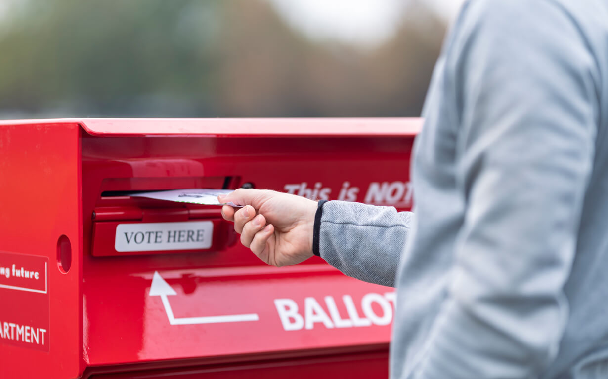 A person, clad in a light gray jacket, places a ballot envelope into a red drop box boldly labeled "Vote Here" and "Ballot."