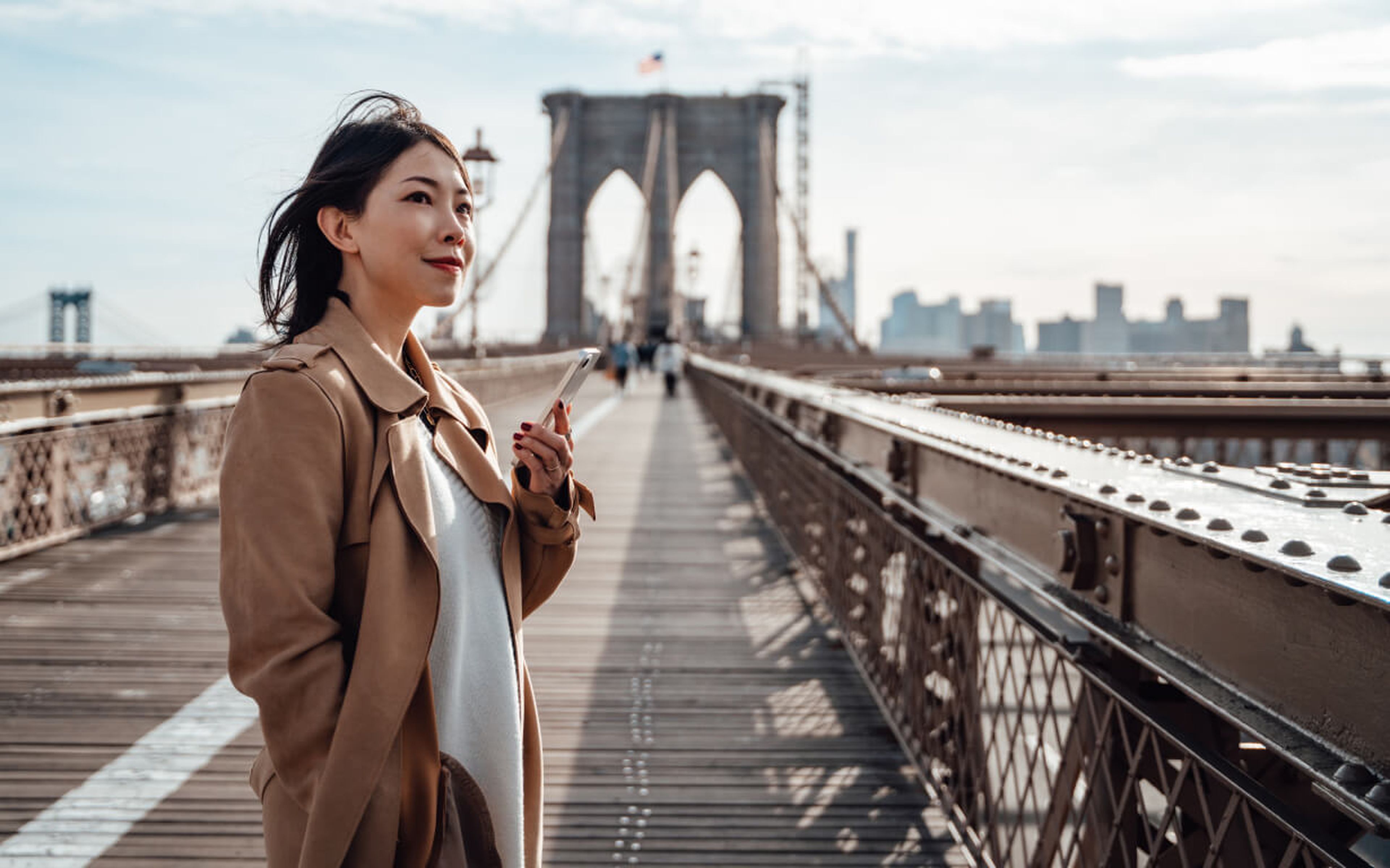 A woman in a tan coat stands on the Brooklyn Bridge, holding her phone and gazing into the distance. The bridge's arches and cables frame the city skyline beneath a partly cloudy sky.
