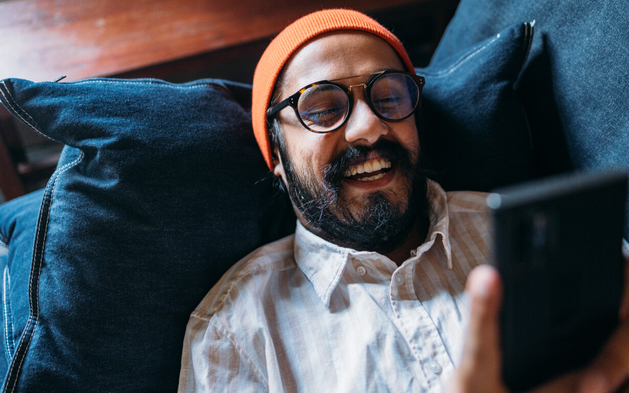 A person with glasses and a beard, wearing a red beanie and a white shirt, is lying on a sofa. They're smiling at their smartphone, possibly exploring how to use a digital wallet, surrounded by blue cushions.