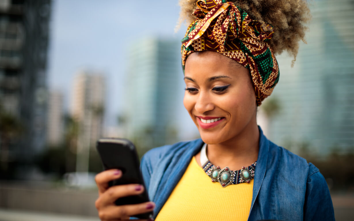 A woman with curly hair and a colorful headwrap smiles while looking at her smartphone. She is outdoors, wearing a denim shirt over a yellow top and a statement necklace. Blurry buildings and palm trees are visible in the background.