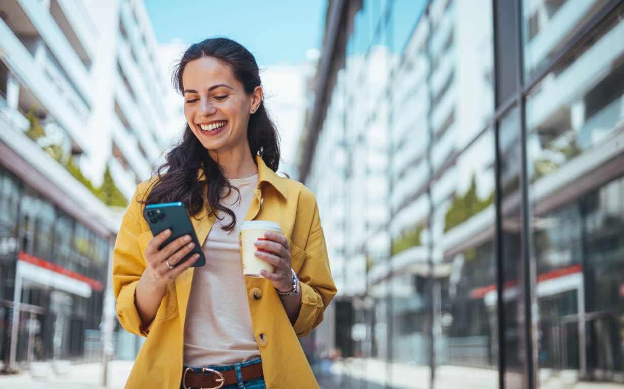 A woman in a yellow jacket smiles while exploring the best ways to send money on her phone. She holds a coffee cup and stands outside between modern glass buildings, enjoying a bright, sunny day in the urban landscape.