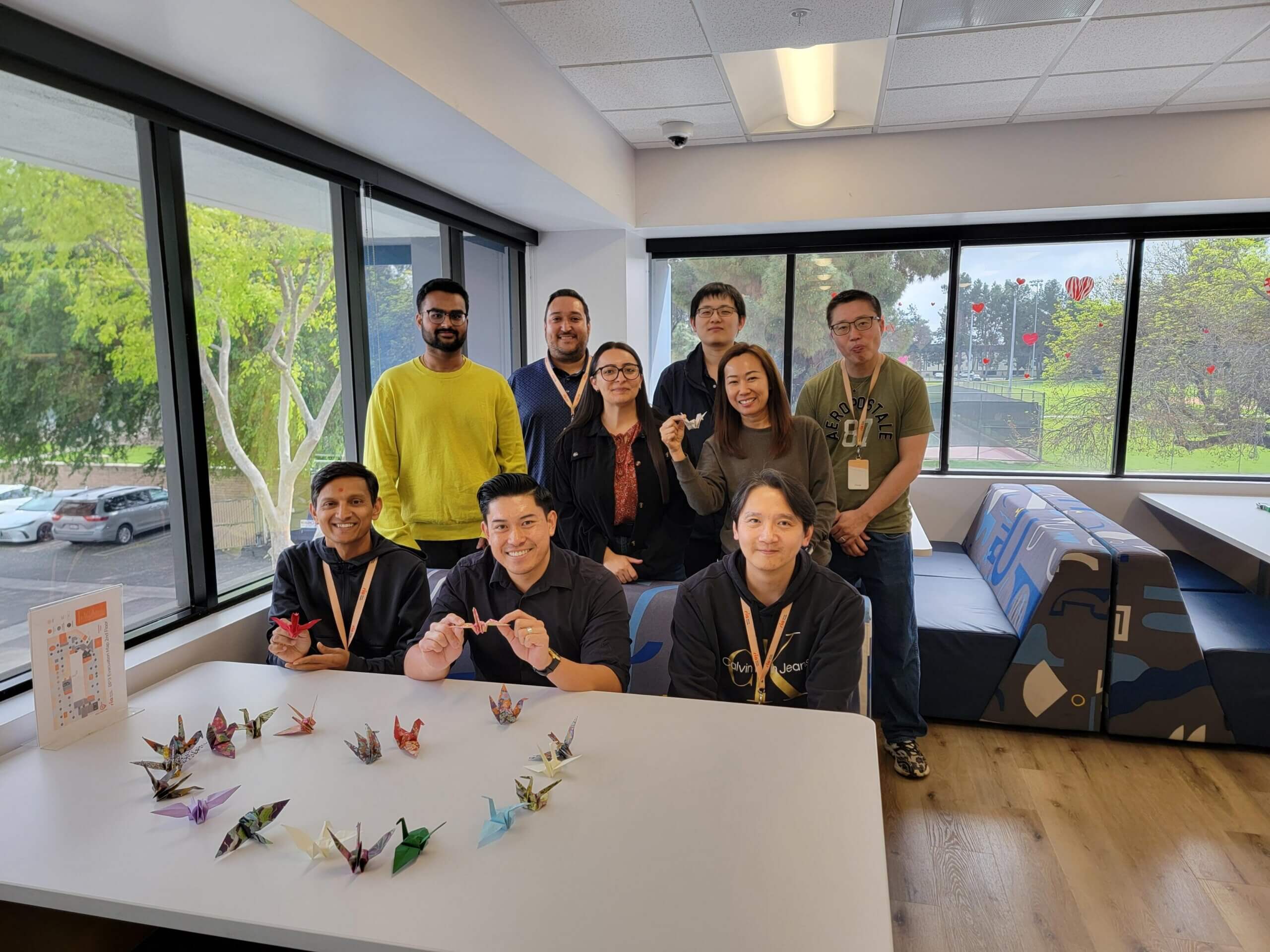 A diverse group of nine people smiles around a table filled with colorful origami cranes.