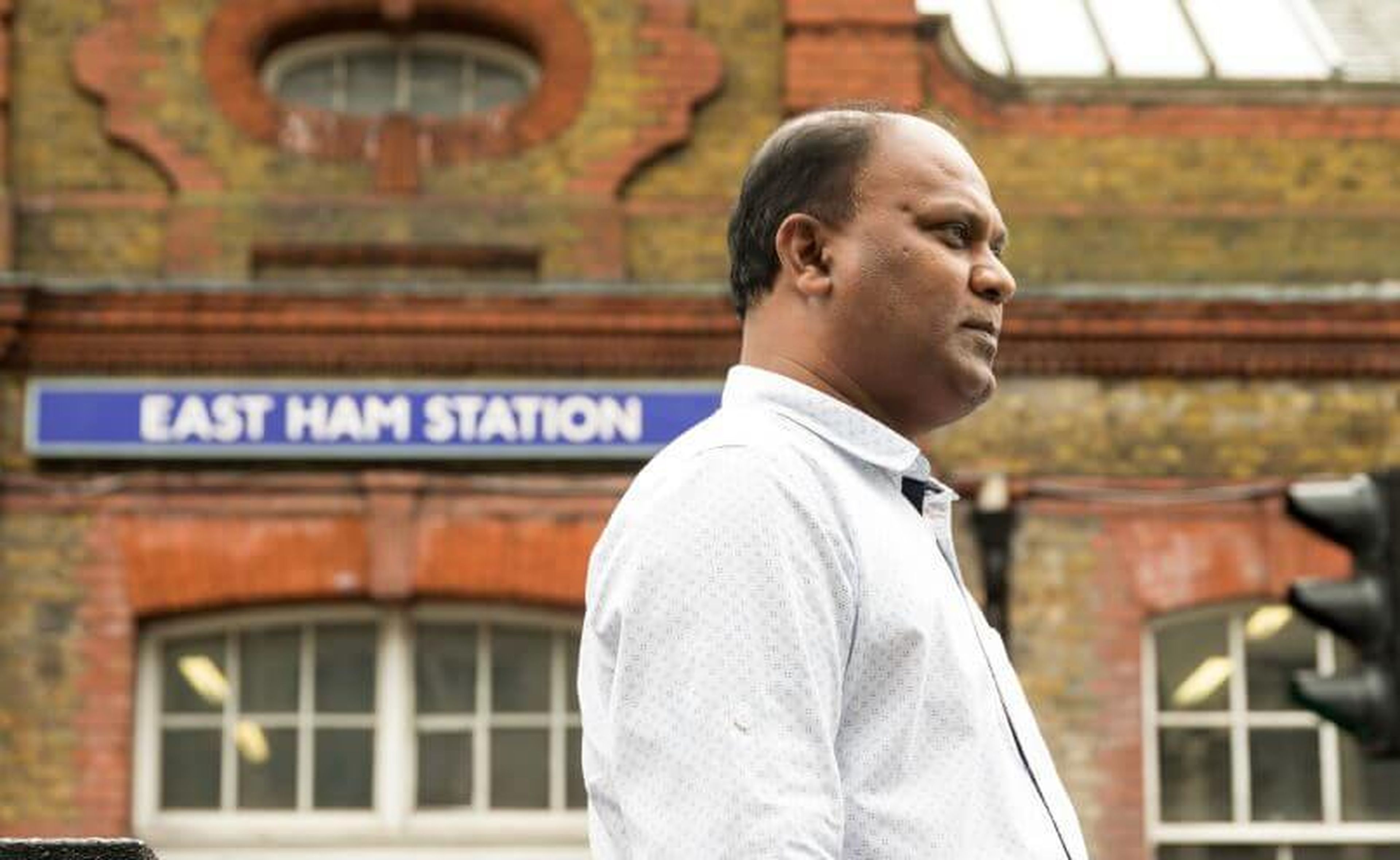 A person, possibly Muhammed, stands in front of East Ham Station, wearing a light-colored shirt. The background showcases the brick facade of the station building with the blue "East Ham Station" sign above.