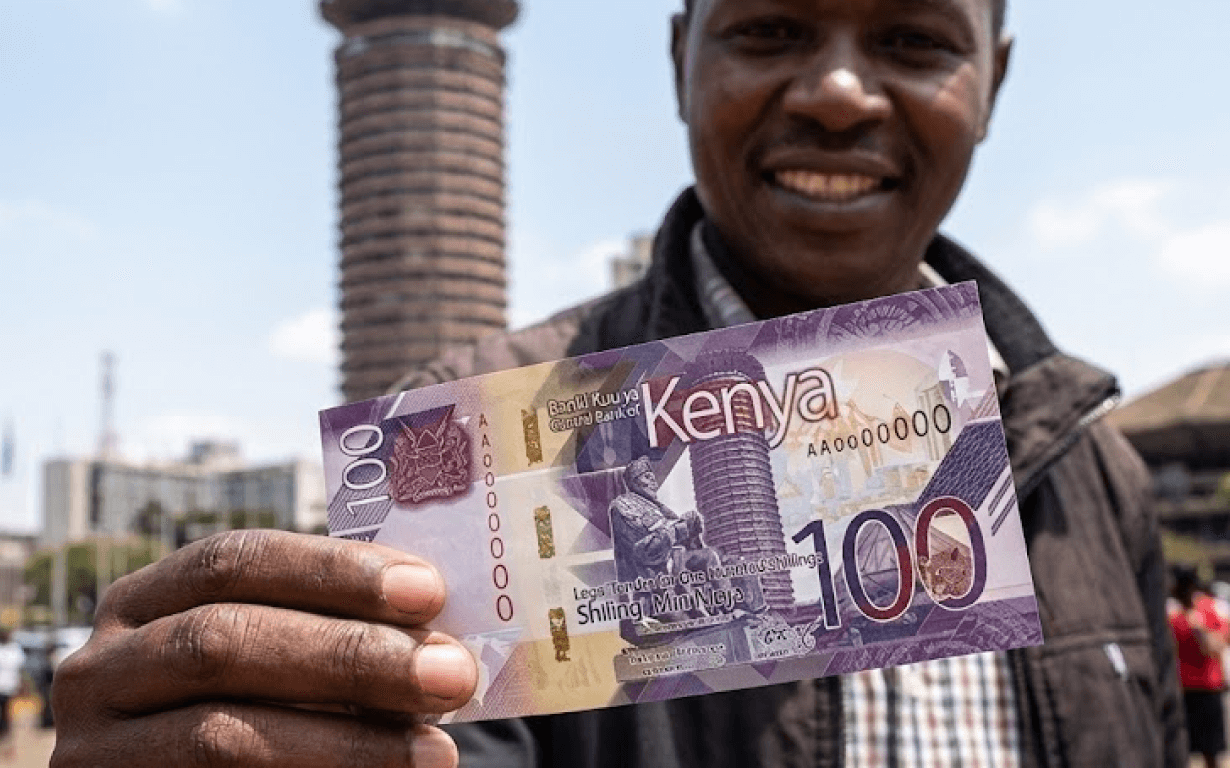A smiling man holds up Kenyan currency—a 100 shilling banknote—in front of tall buildings in an urban outdoor setting. The banknote is prominently displayed, showcasing its design and denomination.