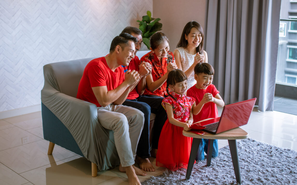 A joyful family of six, including two children and four adults, sits together in a living room, smiling at a laptop. Dressed in red and celebrating the Lunar New Year, they are surrounded by a comfortable setting that enhances their festive spirit.