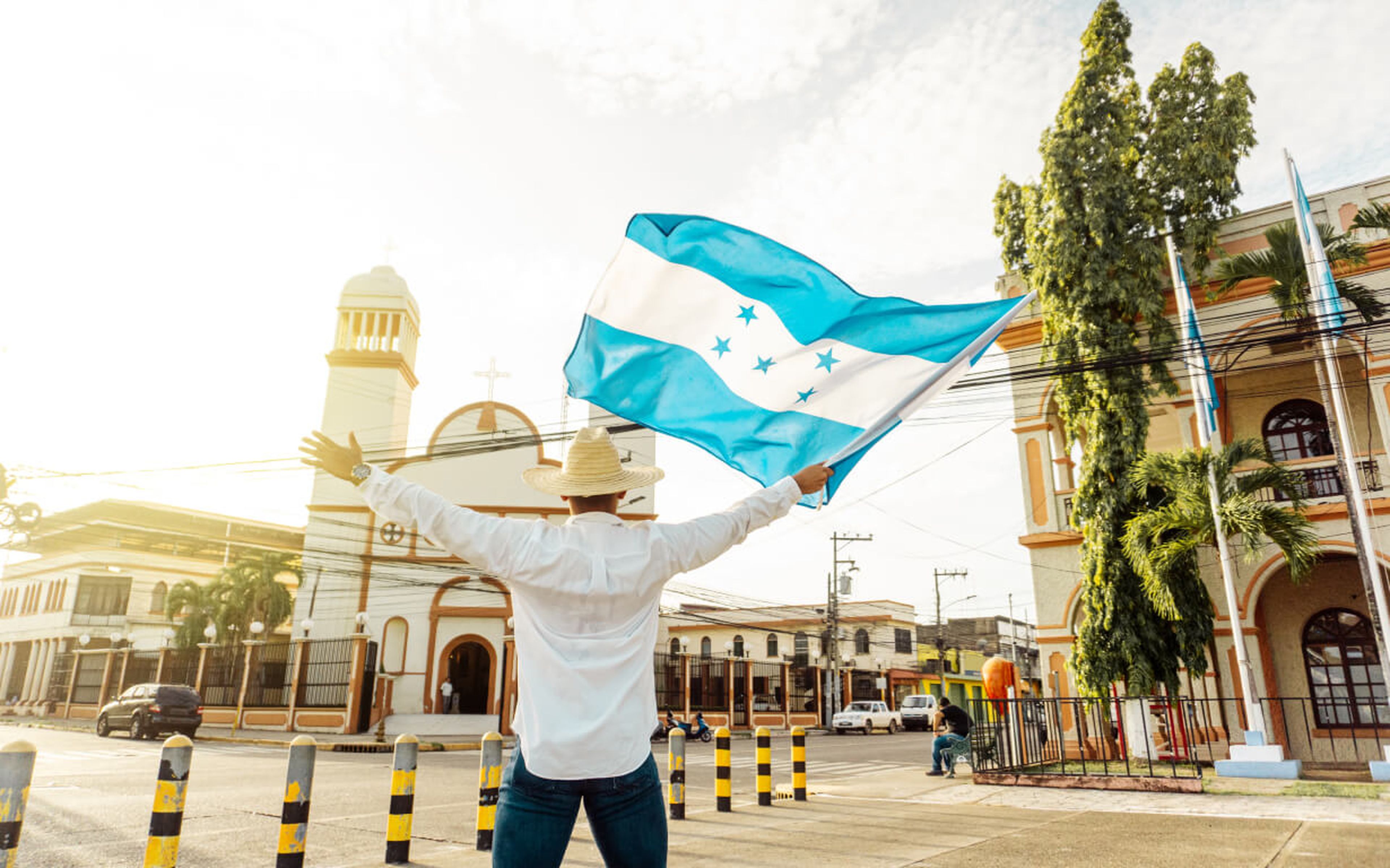 A person standing with arms outstretched, waving the flag of Honduras, wearing a white shirt and a straw hat, celebrates September 15 in front of a colonial-style building and church in a sunlit town square.