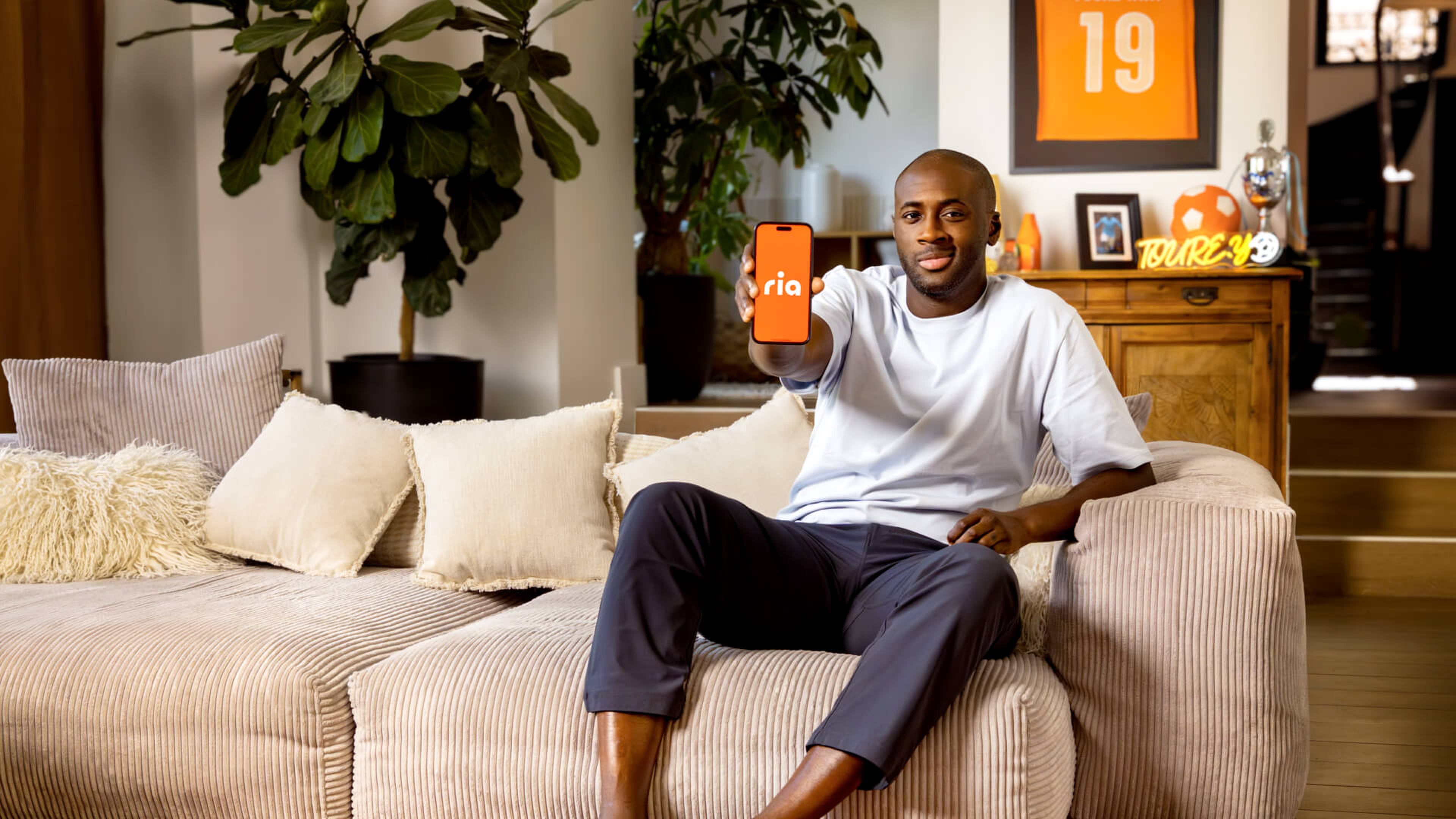 A man sitting on a beige sofa in a cozy living room holds out a smartphone with an orange "ria" logo, smiling at the camera like Yaya Touré. Framed pictures and a plant decorate the background.