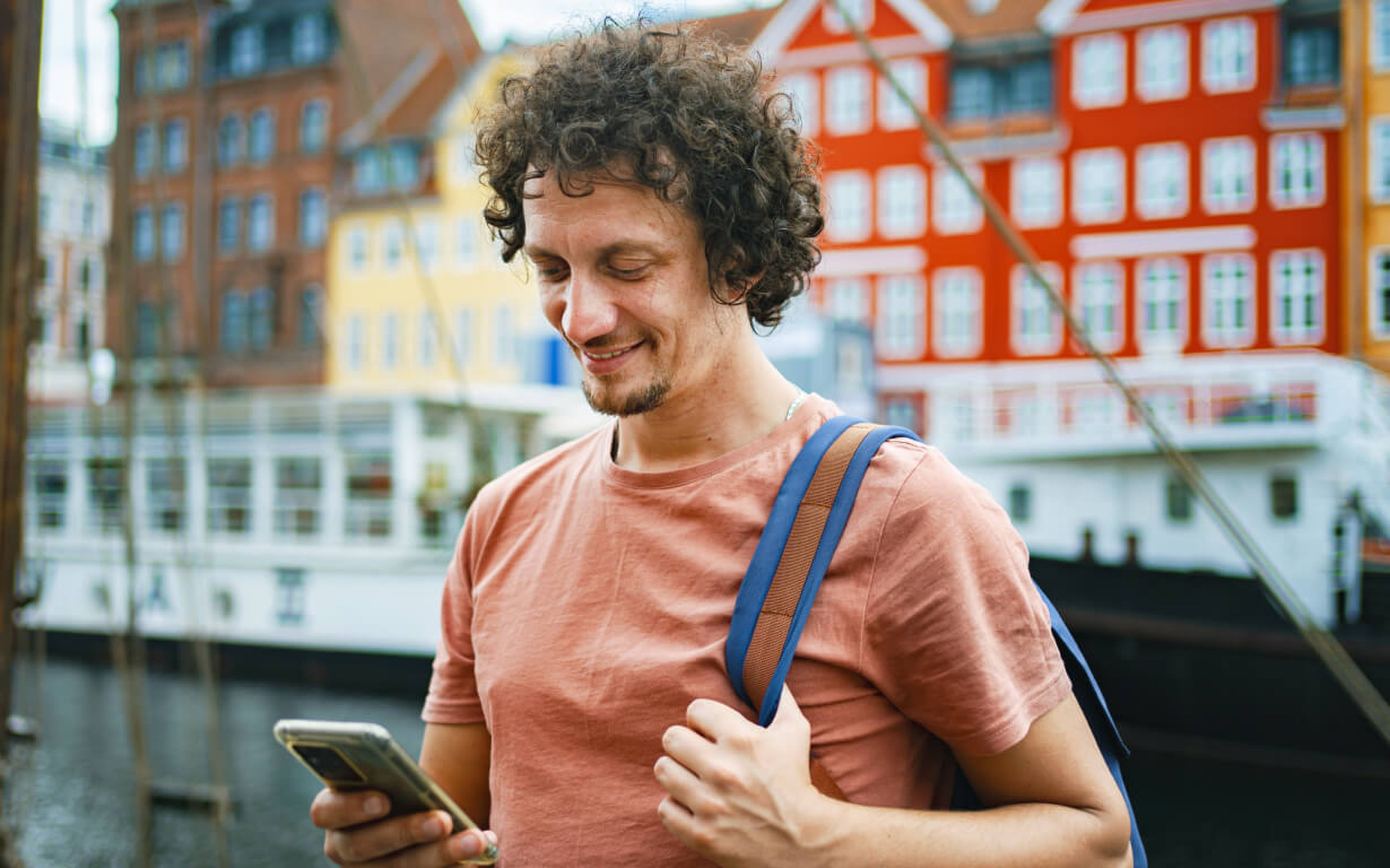 A man with curly hair, wearing a salmon-colored t-shirt and a backpack, smiles while looking at his phone. He stands near a harbor with colorful buildings and boats in the background, perhaps taking a moment to send remittances to loved ones back home.