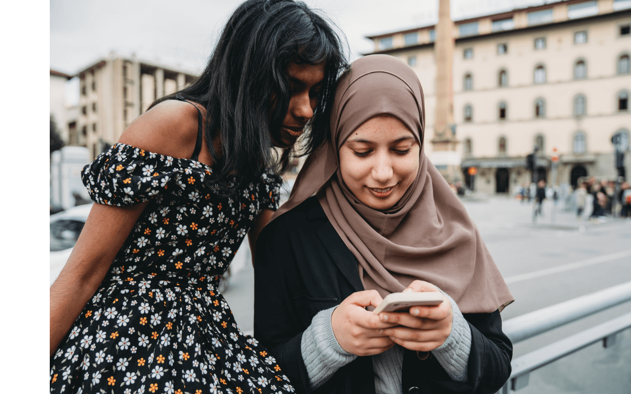 Two women are looking at a smartphone. One has long dark hair and is wearing a floral dress. The other is wearing a light brown hijab and a dark jacket. They are standing outdoors in a public space with buildings in the background, perhaps discussing the Best European Banks to visit on their trip.