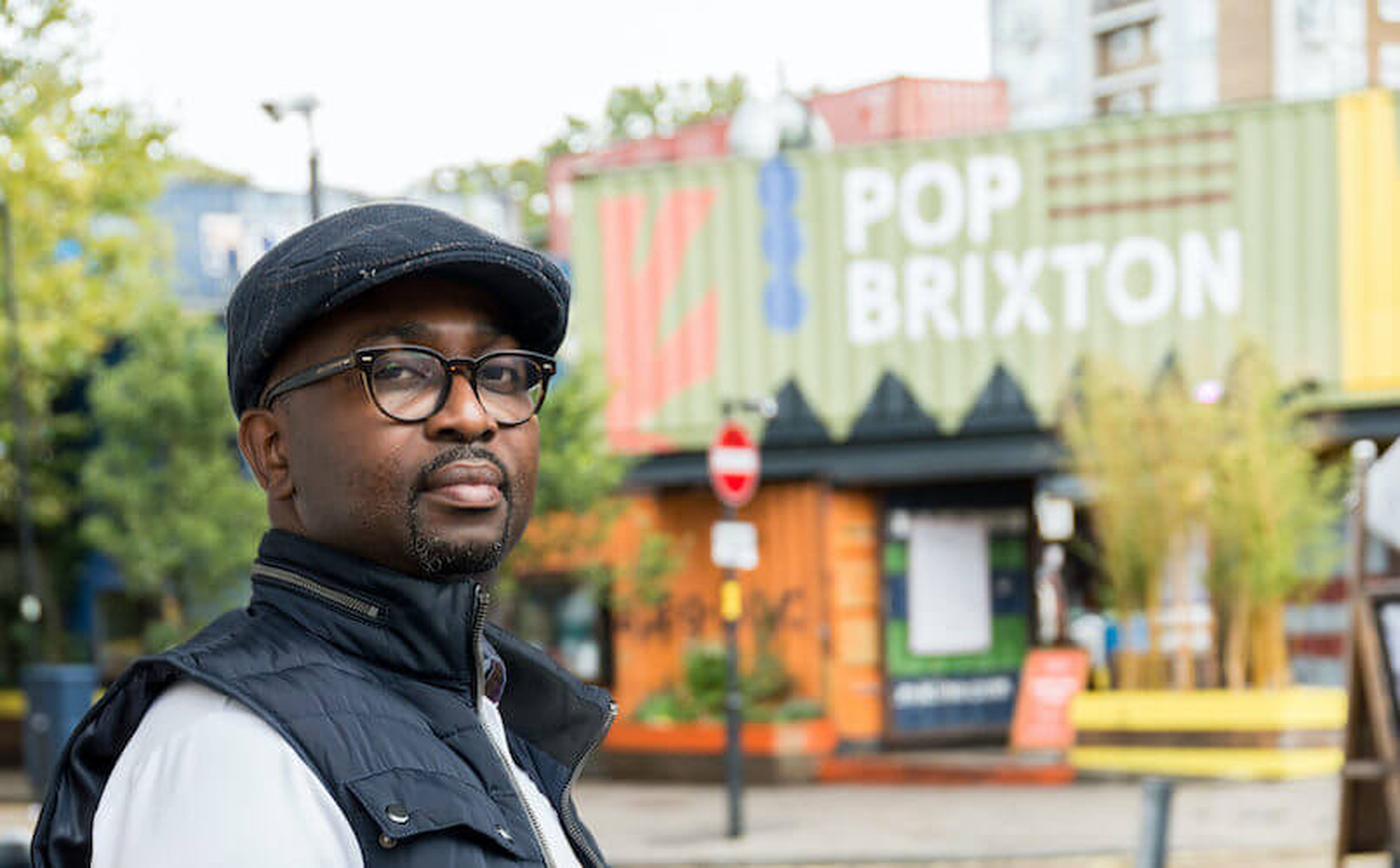 A man wearing glasses and a flat cap stands outdoors, looking at the camera. Behind him is the vibrant public space of "POP BRIXTON," crafted from repurposed shipping containers. Trees and various buildings fill the background, reminiscent of market spaces in the Ivory Coast.