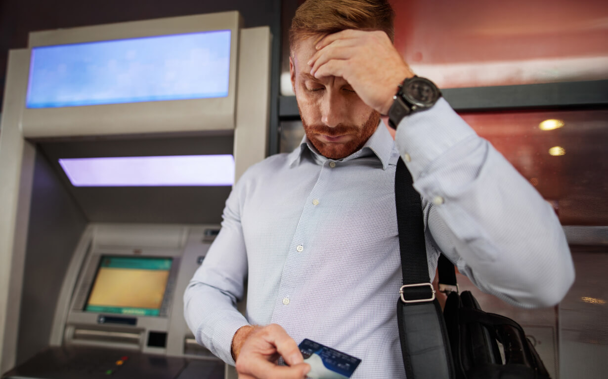 A man standing in front of an ATM looks concerned as he examines a credit card. He is wearing a light blue shirt and a wristwatch, with a bag slung over his shoulder. The ATM screen is blurred in the background.
