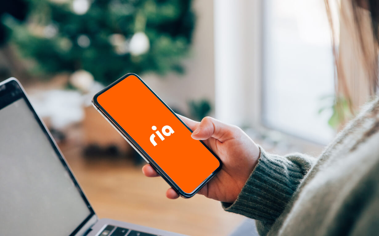 A person holds a smartphone displaying an orange screen with the white text "ria," likely accessing a currency transfer app. They are seated, with part of a laptop visible in the foreground, set against a blurred background of a window and greenery.