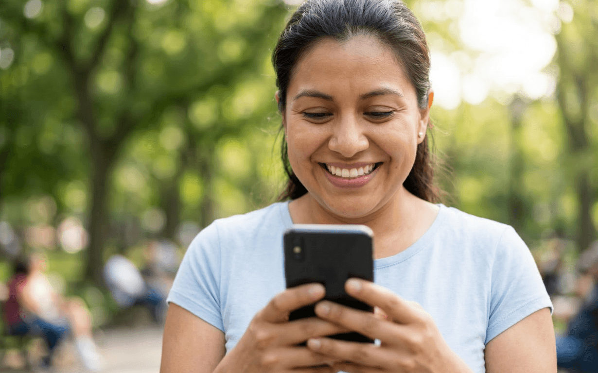 A woman smiles while looking at her smartphone outdoors in a park, happy and engaged as she prepares to Send Money to El Salvador. She wears a light blue shirt, with trees and people blurred in the background.