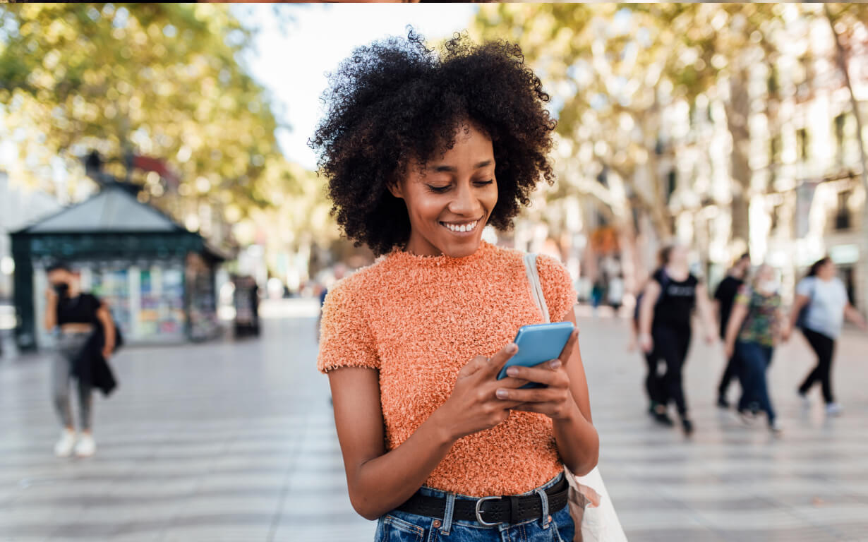 A woman with curly hair wearing an orange sweater smiles while looking at her phone, exploring how to use a digital wallet. She stands on a bustling street lined with trees, as people walk by in the background.
