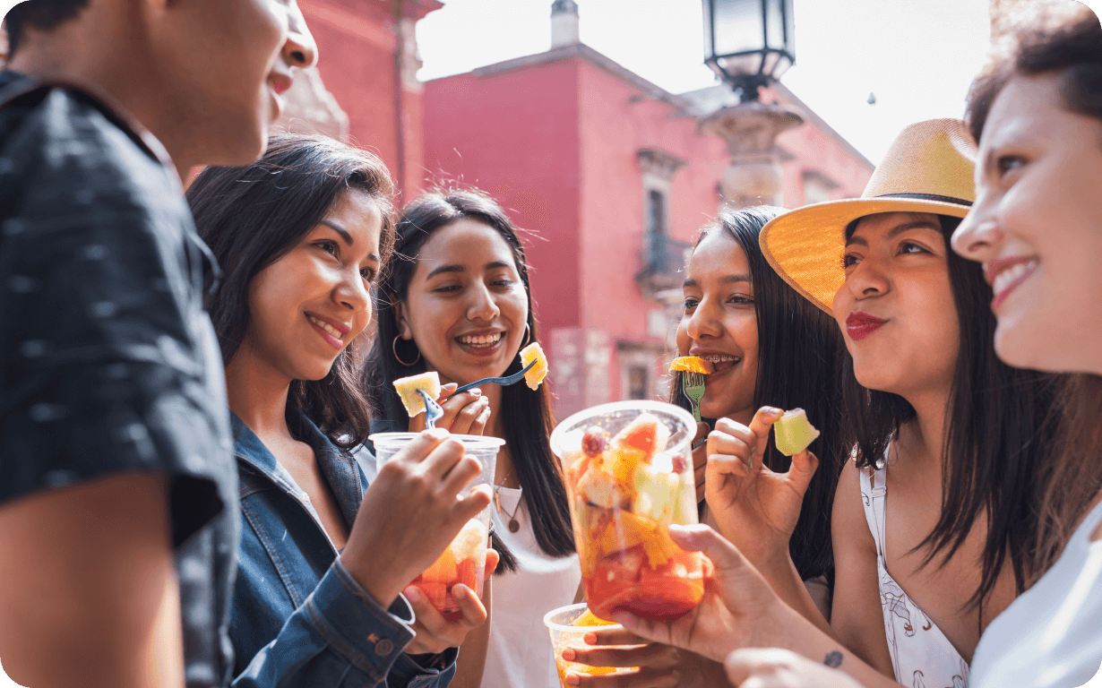 A group of five friends stand together outdoors, smiling and enjoying cups of fresh fruit. The scene is bright and sunny, with colorful buildings in the background. One person wears a straw hat, and everyone appears relaxed and happy, celebrating their recent move to Mexico.