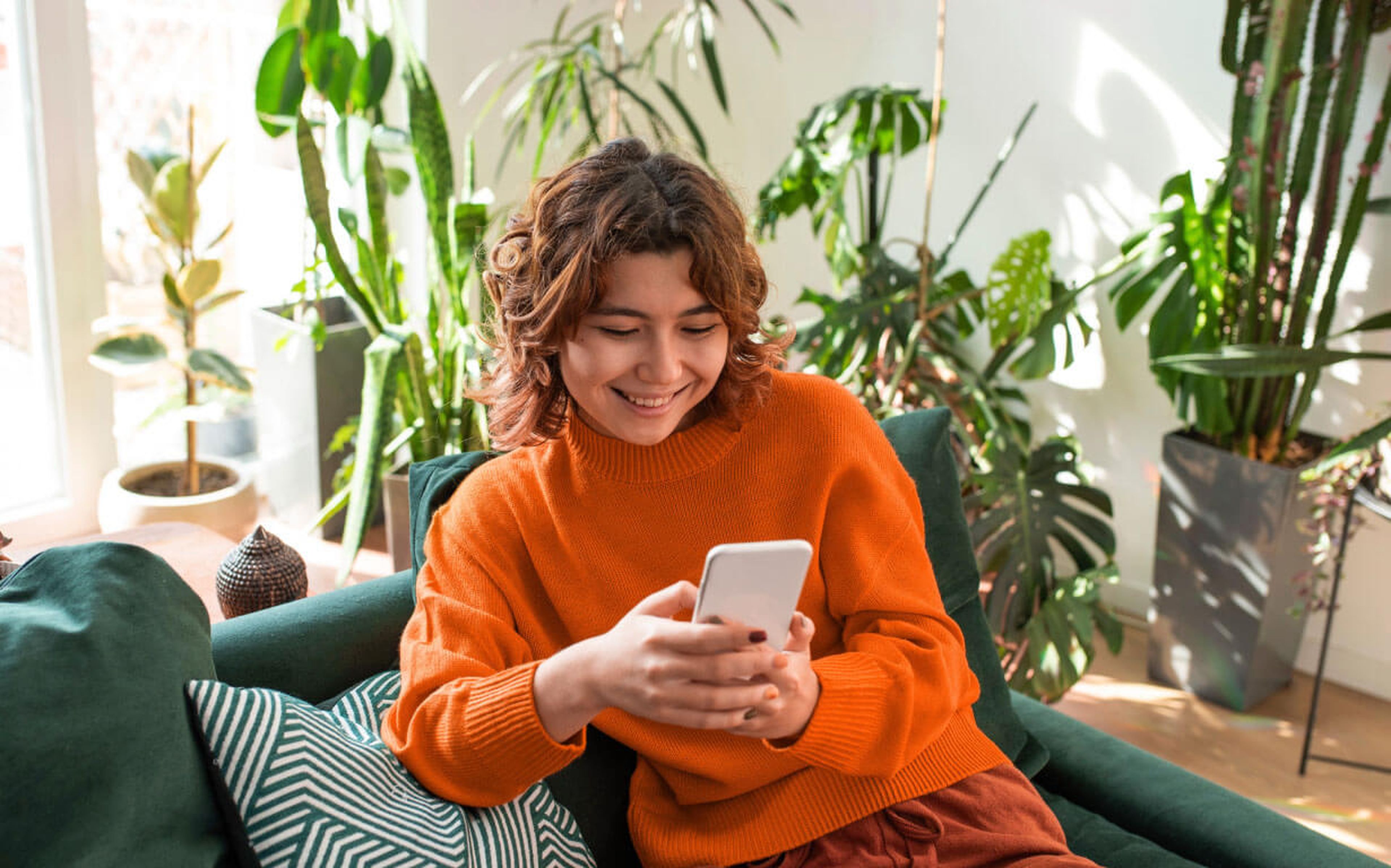 A person with curly hair in an orange sweater is smiling while looking at a smartphone, possibly exploring how to use a digital wallet. They sit on a sofa with green cushions in a plant-filled room, as sunlight filters through the window.