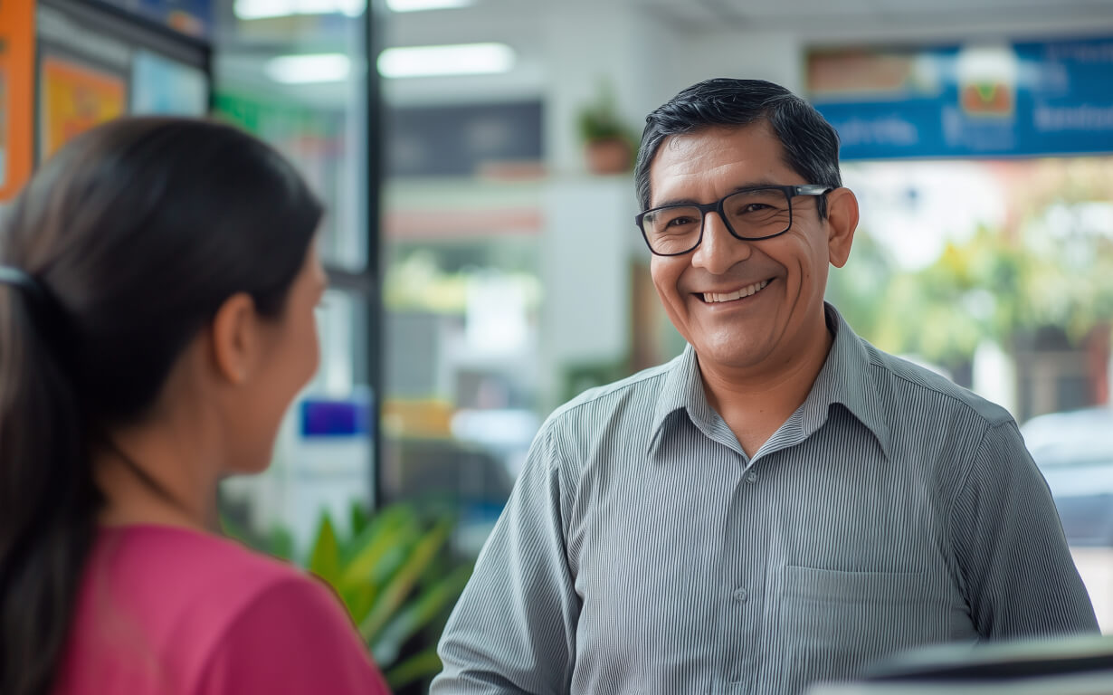 A smiling man wearing glasses and a gray shirt discusses ways to receive money from overseas in the US with a woman in a pink top. They are indoors, possibly in an office or business setting, with plants and blurred signs in the background.