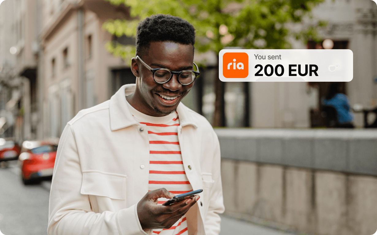 A smiling man wearing glasses and a white jacket looks at his phone outdoors. A notification on the image shows he sent 200 EUR using the Ria money transfer app.