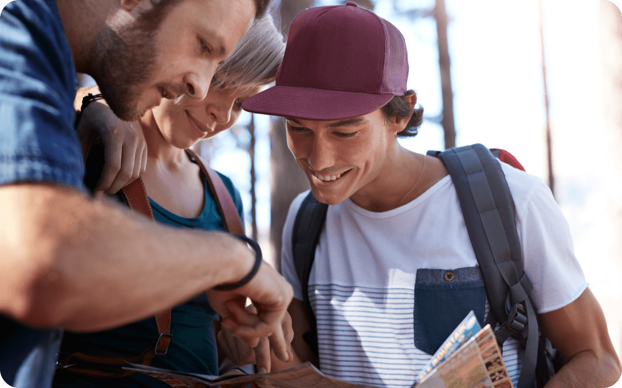 Three friends, wearing casual outdoor clothing and backpacks, are looking at a map together. One person is pointing at the map while the other two smile, with trees visible in the background. They seem to be enjoying a hike during their gap year abroad, embracing their outdoor adventure.