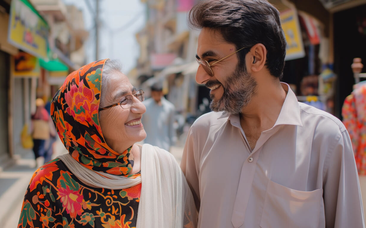 A smiling elderly woman in a colorful scarf and a man in glasses and a light shirt walk closely together on a sunny street, warmly looking at each other. The background features blurred shops and people from vibrant migrant communities.