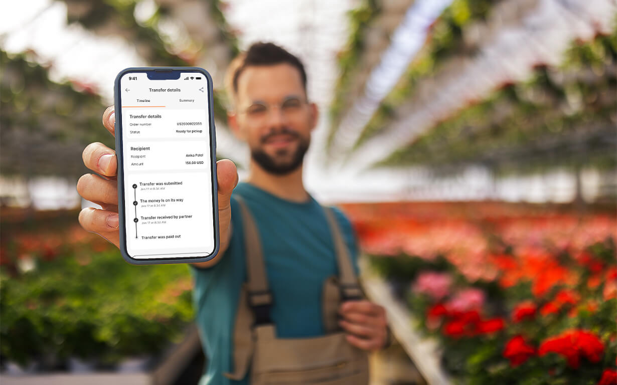 A person wearing glasses and overalls stands in a greenhouse, holding a smartphone towards the camera with the ria app open to a bank transfer details page. Rows of flowers and plants are visible in the background.