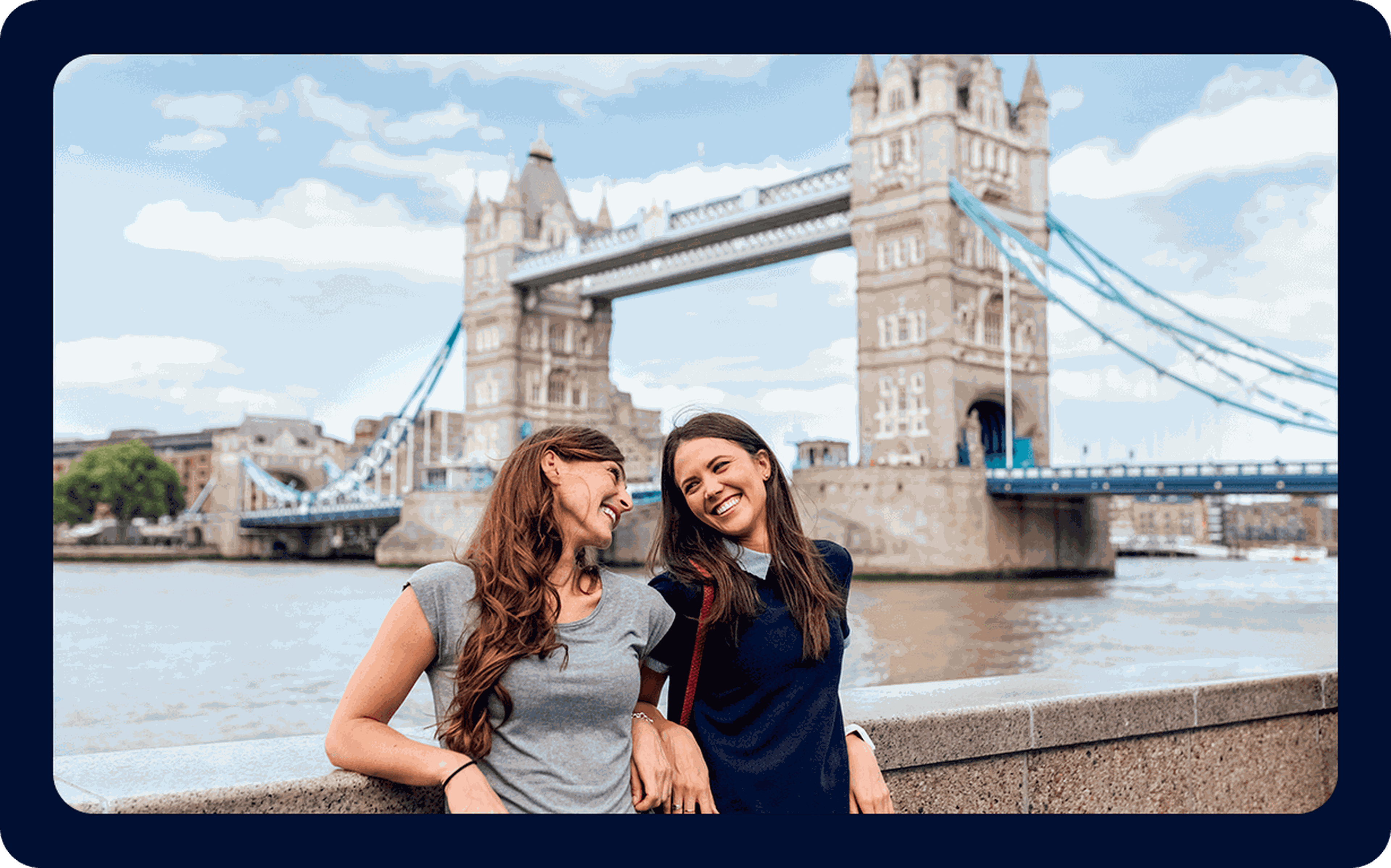 Two smiling women stand in front of the Tower Bridge in London, leaning on a stone railing as they enjoy the view and each other's company. The sky is partly cloudy, and the iconic bridge forms a picturesque background—a perfect moment for celebrating their decision about moving to the UK.