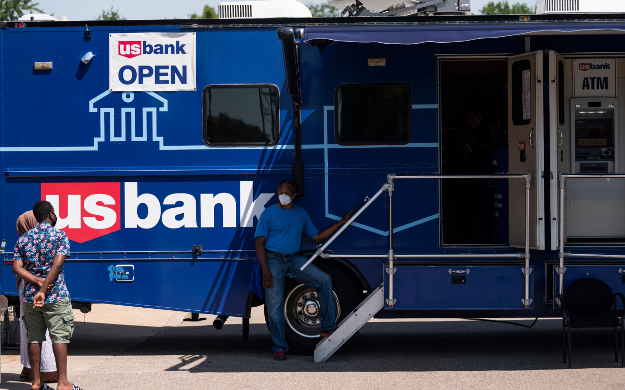 A person stands outside a blue mobile banking vehicle with "US Bank OPEN" signage, one of the 5 Best US Banks. An ATM is visible inside. Another person is nearby, facing the vehicle under the bright sun on this cheerful day.