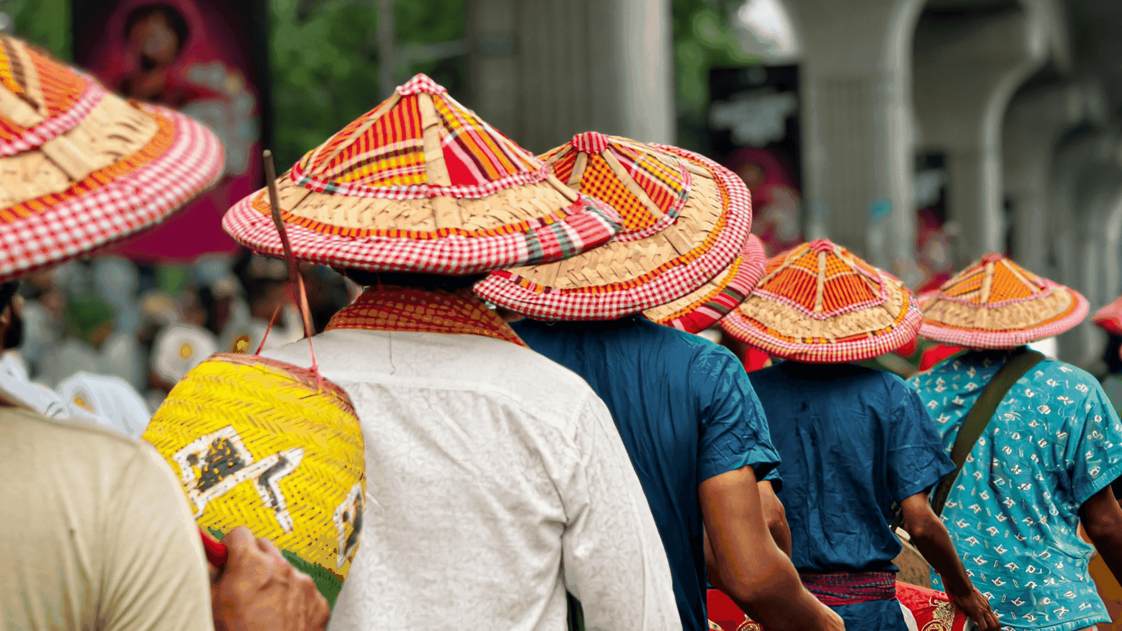 A group of people wearing colorful, traditional woven hats and carrying baskets walk in a line outdoors, with greenery and columns visible in the background.