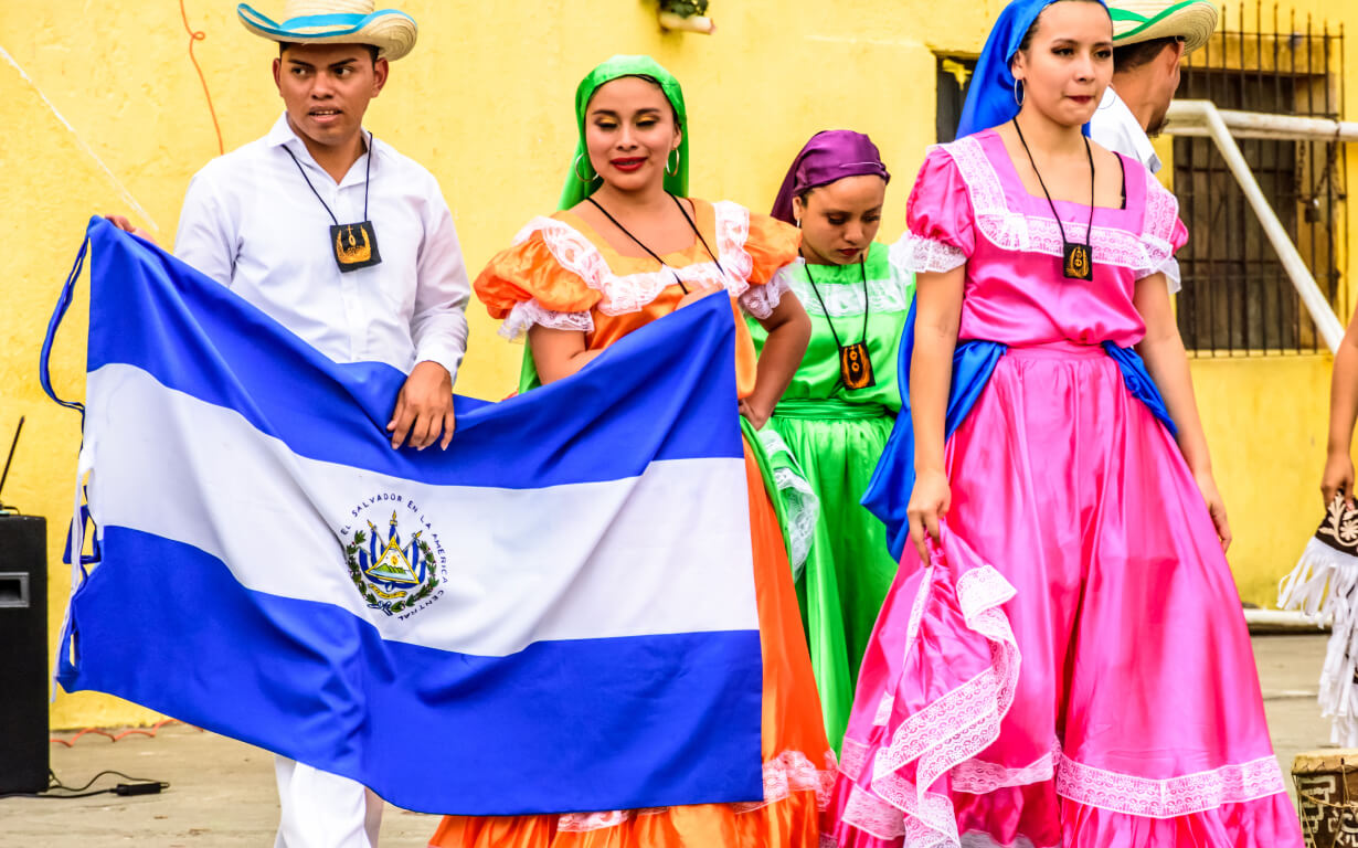 A group of people in colorful traditional clothing stand together. One of them holds a blue and white flag with a coat of arms in the center, likely for a September 15 celebration. The background features a yellow wall. The attire includes vibrant dresses and hats, suggesting a cultural celebration.