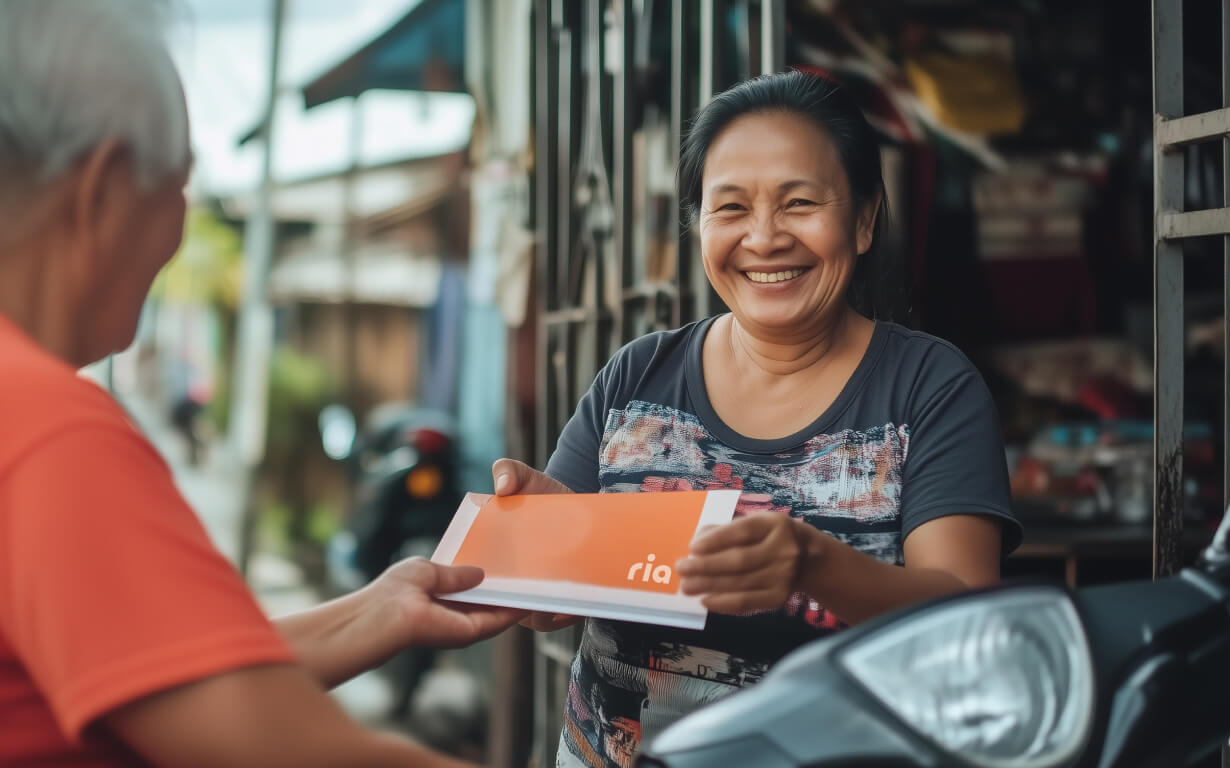 A smiling woman hands an orange and white package to another person in an outdoor setting, reminiscent of the seamless ease when you receive money from overseas in the US. She stands near a building with blurred backgrounds featuring a street and motorcycle.
