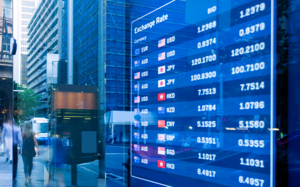 A digital screen showcases currency exchange rates against the U.S. dollar, highlighting the euro, yen, pound, and more. The reflection of people walking and urban buildings appears on the sleek glass surface.